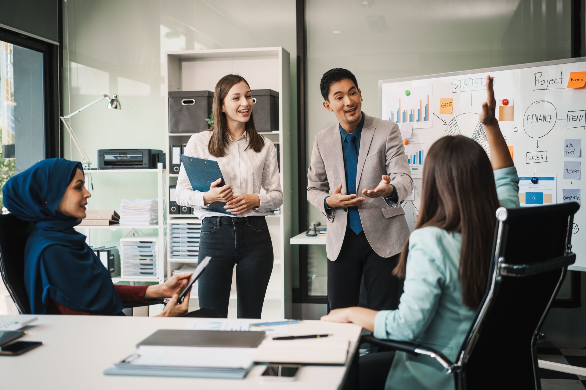 A diverse team, including a middle-aged Asian businessman, Caucasian young businesswoman, Muslim hijab-wearing middle-aged businesswoman, meets in a boardroom to close an Annual General Meeting.